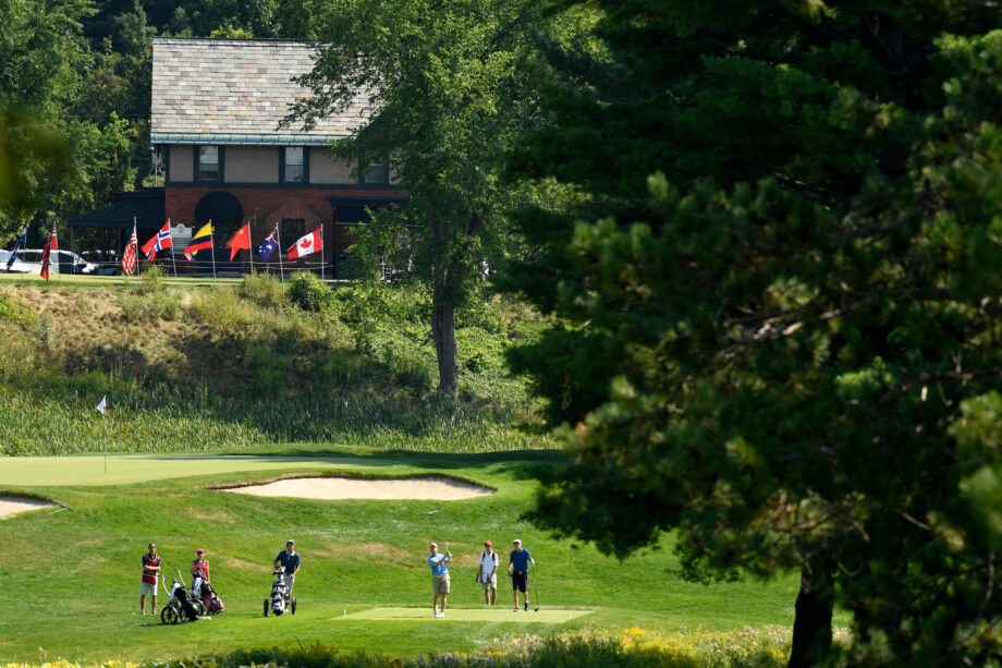 Un groupe de golfeurs se tient sur un départ verdoyant au Club de golf Royal Ottawa, avec des drapeaux internationaux flottant en arrière-plan devant un bâtiment en briques.