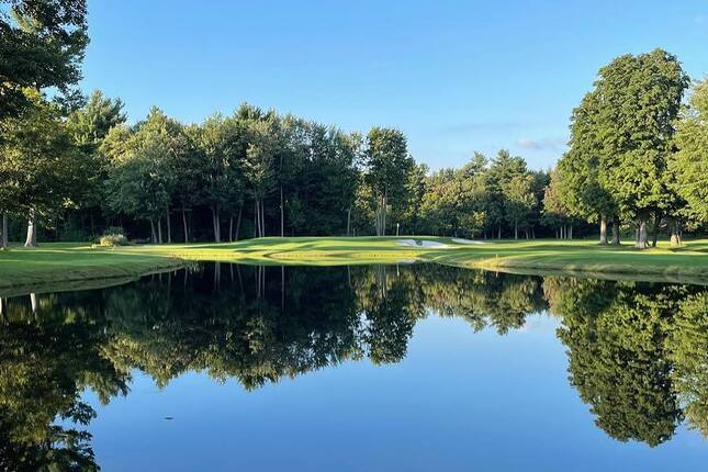 Un lac calme reflète le terrain de golf verdoyant de la Vallée du Richelieu, bordé d’arbres sous un ciel bleu clair.
